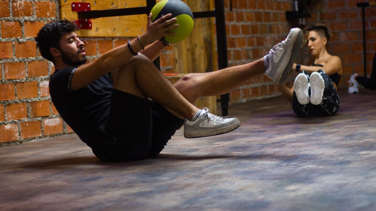 A man and woman perform core exercises with a medicine ball in a brick wall gym.